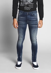 Jeans skinny de color azul oscuro con desgastes, pasadores para cinturón y un diseño de cinco bolsillos. Combinados con una camiseta negra de manga larga y zapatillas marinas.