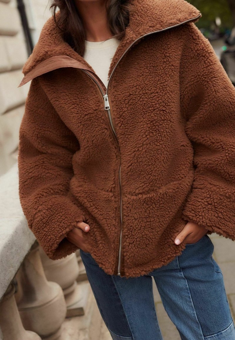 Veste marron en tissu duveteux avec fermeture éclair portée sur un haut blanc, mains dans les poches, associée à un jean patchwork bleu à l'extérieur près d'une rambarde en pierre.