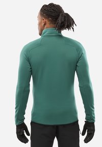 Langärmliges, grünes Fleece-Half-Zip-Oberteil mit glatter Textur, hoher Kragen und enganliegenden Ärmeln, kombiniert mit schwarzen Handschuhen.