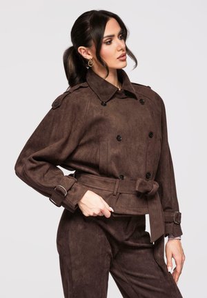 Kvinde med mørk hestehale iført en brun dobbeltradet jakke med bælte og matchende bukser, poserer med den ene hånd på hoften.