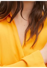 Gelbe Wickelbluse mit langen Ärmeln, V-Ausschnitt und weichem Stoff. Verfügt über eine glatte Textur und fällt an den Schultern elegant.