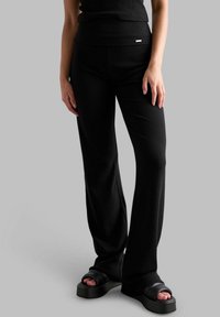 Schwarze, gerippte Strickhosen mit hoher Taille und weit ausgestellten Beinen. Kombiniert mit klobigen schwarzen Sandalen. Der Hintergrund ist schlicht grau.