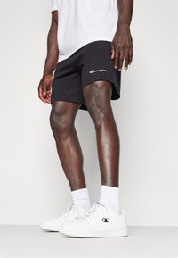 Black shorts with logo, cotton blend fabric, side pockets, and elastic waistband. Paired with white sneakers and crew socks.