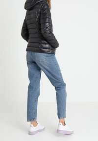Person som bär en svart puffjacka, ljusblå jeans och vita sneakers med lila sulor, står mot en enfärgad ljus bakgrund.