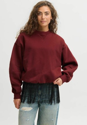 Une femme porte un sweat oversize bordeaux à franges noires, associé à un jean clair déchiré, sur un fond de studio uni.