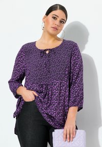 Lila Blumenbluse mit Dreiviertelärmeln, strukturiertem Stoff, rundem Ausschnitt und Bindedetail am Kragen; kombiniert mit dunkelblauen Jeans und einer hellen Handtasche.