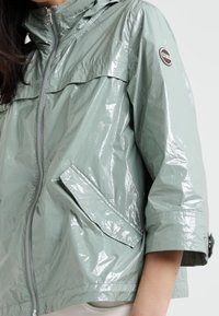 Světlá zelená, lesklá dešťová bunda se zipem vpředu, polovičními rukávy a bočními kapsami. Obsahuje logo na levém rameni a kapuci.