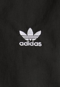 Czarna tkanina z białym haftowanym logo Adidas w kształcie trifolii oraz trzema poziomymi paskami poniżej, podkreślająca gładką teksturę.
