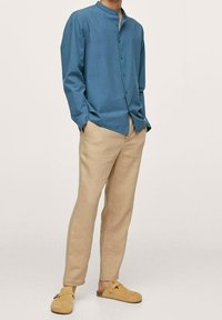 Camicia azzurra a maniche lunghe con colletto alla cinese, pantaloni beige e scarpe slip-on in camoscio marroni, con una vestibilità rilassata e una texture leggera.