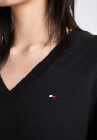 Svart strikket V-hals genser med et lite logo-patch i rødt, hvitt og blått på venstre bryst. Myk tekstur med en tettsittende passform.