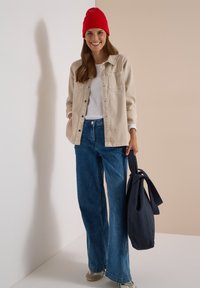 Jeune femme souriante portant un bonnet rouge, une veste beige, une chemise blanche, un jean large bleu, des chaussures grises, tenant un grand sac bleu foncé.