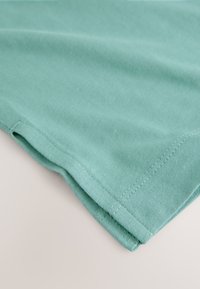 Material de bumbac verde deschis cu o textură netedă, având o margine pliată și cusături contrastante de-a lungul marginii.