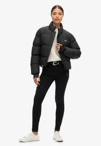 Zwarte pufferjack met hoge kraag, ritssluiting aan de voorkant en geribde manchetten, gedragen over een crèmekleurige trui en gecombineerd met zwarte jeans en witte sneakers.
