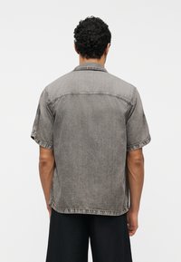 Άνδρας με σγουρά μαλλιά που φοράει κοντομάνικο ξεβαμμένο γκρι τζιν πουκάμισο και μαύρο παντελόνι, στέκεται αντιμετωπίζοντας ένα απλό λευκό φόντο.