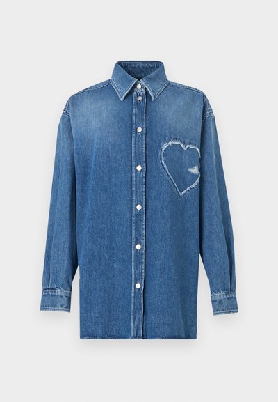 Chemise en denim bleu à manches longues, boutonnée, avec un col pointu et un patch en forme de cœur effiloché sur le côté gauche de la poitrine.
