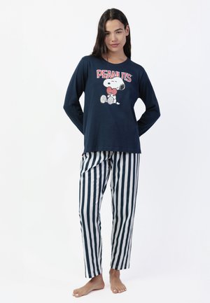 Mujer de pie descalza, vistiendo una camiseta de manga larga azul marino con Snoopy y pantalones de pijama a rayas negros y blancos sobre fondo blanco.