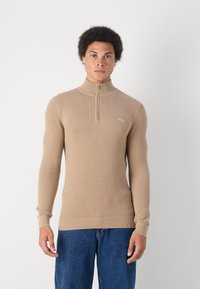 Maglione con zip beige, caratterizzato da un design a maglia testurizzata, maniche lunghe aderenti e orlo a coste. Un logo discreto è visibile sul petto.