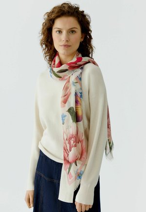 Woman with curly hair wearing a white sweater, dark skirt, and a light floral scarf draped around her neck, standing against a plain background.