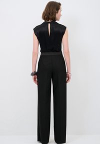 Top sans manches en satin noir avec un dos en trou de serrure, associé à un pantalon noir ajusté taille haute. Bracelets transparents visibles aux poignets.