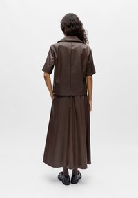 Femme portant une chemise à manches courtes en similicuir marron et une jupe longue assortie, avec des chaussettes foncées et des mocassins noirs, debout devant un fond blanc.