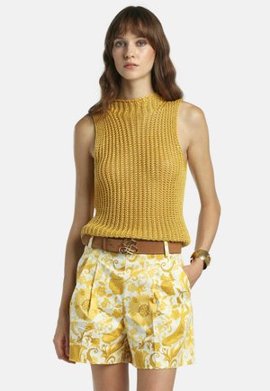 Žena v bezrukávovom horčicovom pletenom topu s vysokým golierom, žltých šortkách s kvetinovým vzorom, hnedom opasku a zlatom náramku, stojaca s jednou rukou v vrecku.