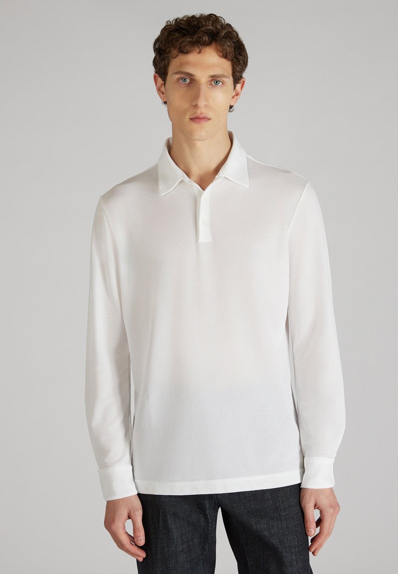 Witte lange mouw polo met een subtiele textuur, knoopsluiting en contrasterende kraag. Gedragen met donkere jeans.