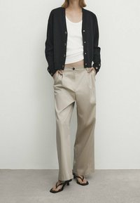 Czarny kardigan z detalami guzika na białej bluzce na ramiączkach, skomponowany z beżowymi spodniami typu wide-leg i czarnymi klapkami na gładkiej powierzchni.