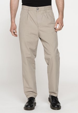 Uomo che indossa pantaloni beige a gamba dritta con pieghe e scarpe nere di pelle con lacci su sfondo semplice.
