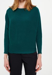 Teal geribbelde trui met een ontspannen pasvorm, ronde halslijn en lange mouwen, in combinatie met zwarte broeken. Het materiaal lijkt zacht en gestructureerd.