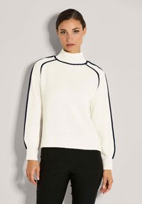 Frau in einem weißen Rollkragenpullover mit schwarzem Piping an Ärmeln und Schultern, kombiniert mit schwarzen Hosen, steht vor einer schlichten Wand.