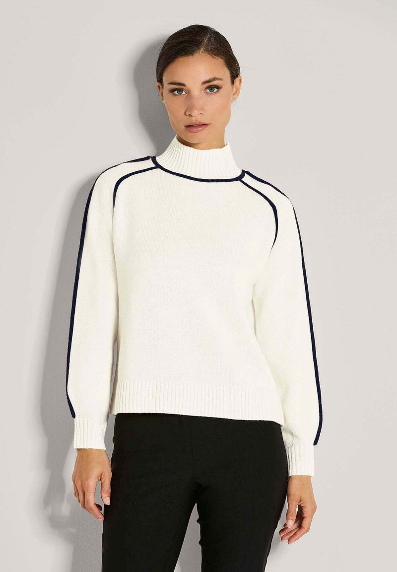 Frau in einem weißen Rollkragenpullover mit schwarzem Piping an Ärmeln und Schultern, kombiniert mit schwarzen Hosen, steht vor einer schlichten Wand.