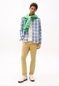 Camisa de cuadros azul y blanca, sudadera verde sobre los hombros, camiseta blanca, pantalones beiges y zapatillas blancas con detalles en negro.