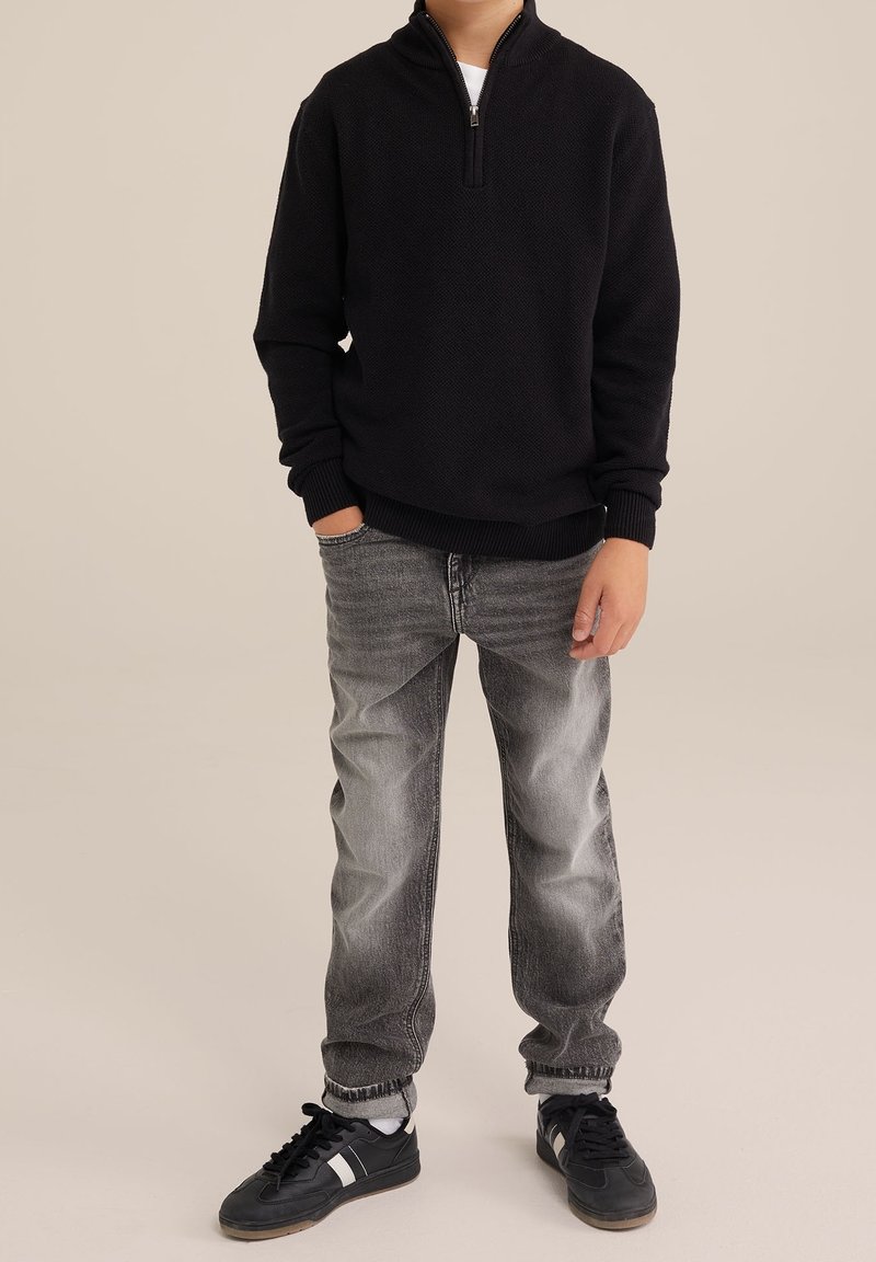 Garçon portant un pull noir à fermeture éclair quart, un jean gris passé et des baskets noires avec des accents blancs, debout avec une main dans la poche.