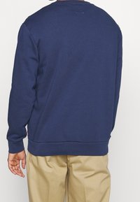 Sudadera azul marino con puños y dobladillo acanalados, hecha de tela suave. Vista trasera muestra una textura lisa y un branding minimalista cerca del dobladillo.