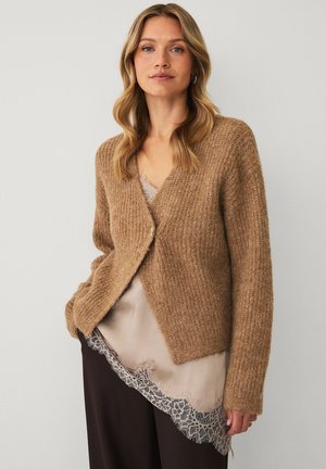 Woman with wavy blonde hair wears a brown knit cardigan over a beige lace-trimmed top and dark wide pants against a plain background.