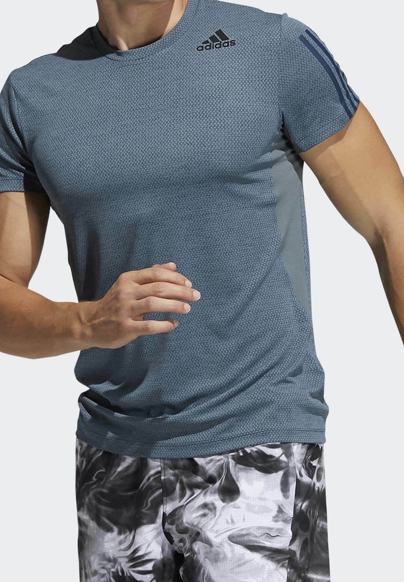 Hombre vistiendo una camiseta de manga corta texturada azul de Adidas y pantalones cortos con patrón en blanco y negro, en movimiento con los brazos flexionados.