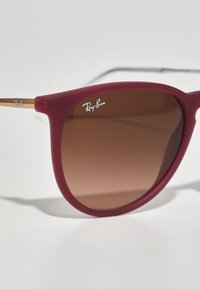 Ochelari de soare Ray-Ban cu un cadru mat de culoare burgundă, lentile cu gradient brown și un accent metalic de aur pe brătara laterală. Logo-ul este afișat pe cadru.