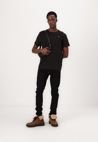 T-shirt noir à col rond, jeans noirs et bottes marron avec un col beige. Accessoire : sac messager noir avec une sangle logo.