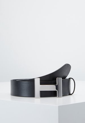 Ceinture en cuir noir avec une grande boucle métallique argentée en forme de la lettre H, présentée enroulée sur une surface blanche.