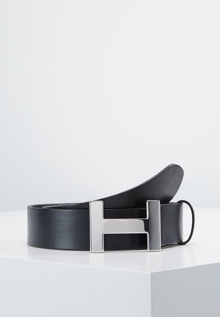 Ceinture en cuir noir avec une grande boucle métallique argentée en forme de la lettre H, présentée enroulée sur une surface blanche.