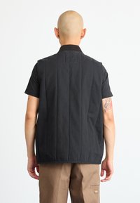 Črn brezrokaven jopič z visokim ovratnikom, teksturirana tkanina in vidne šive, v kombinaciji s črno srajco s kratkimi rokavi in bež hlačami.