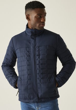 Mężczyzna w granatowej pikowanej kurtce z rękami w kieszeniach, patrzący w bok, ubrany w niebieskie jeansy na jednolitym tle.