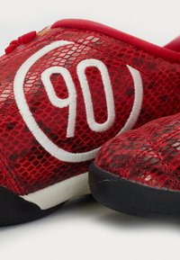 Röda sneakers med överdel i mesh, med en vit broderad "90" logotyp, svarta sulor och ett strukturerat mönster över materialet.