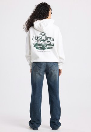 Witte hoodie met groene grafische tekst en klassieke autoafbeelding op de achterkant. Samen gedragen met hoge, losse spijkerbroeken in het blauw.