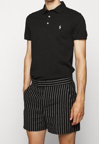 Polo noir avec une patte de boutonnage à trois boutons et un logo blanc, associé à un short noir rayé avec une large ceinture et des poches.