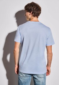 T-shirt bleu clair à manches courtes avec un col rond, texture en coton lisse et ourlets cousus, présenté de dos avec un jean bleu délavé.