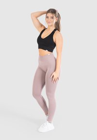 Reggiseno sportivo nero a coste con scollatura a V e leggings a vita alta di un delicato color malva. Modella che indossa sneakers bianche, in posa con una mano sulla testa.