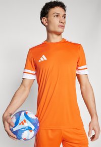 Orange athletic jersey with white stripes on sleeves, featuring the Adidas logo. The model holds a blue and white soccer ball.