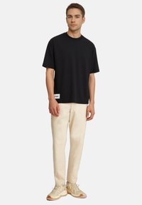 Maglietta nera in cotone con collo rotondo e maniche corte, abbinata a pantaloni beige chiaro. Presenta un'etichetta di marca sull'orlo.