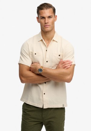 Homme bras croisés portant une chemise beige à manches courtes avec boutons, un pantalon vert olive et une montre-bracelet avec un bracelet beige.